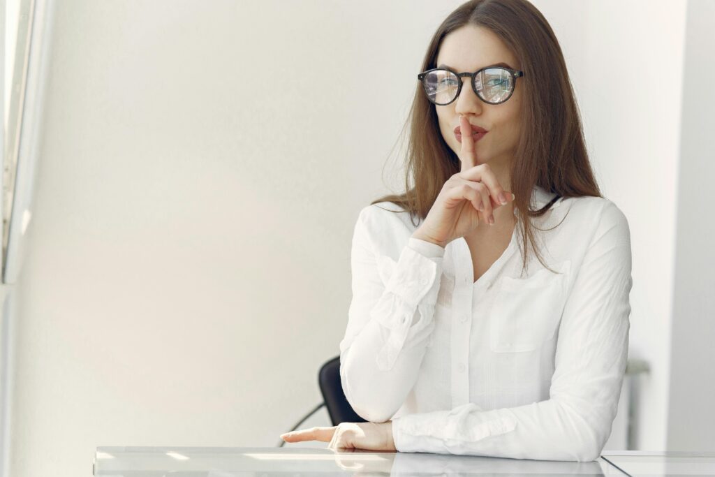 Young woman with long hair in trendy clothes sitting at table and doing shh gesture asking to stay quiet