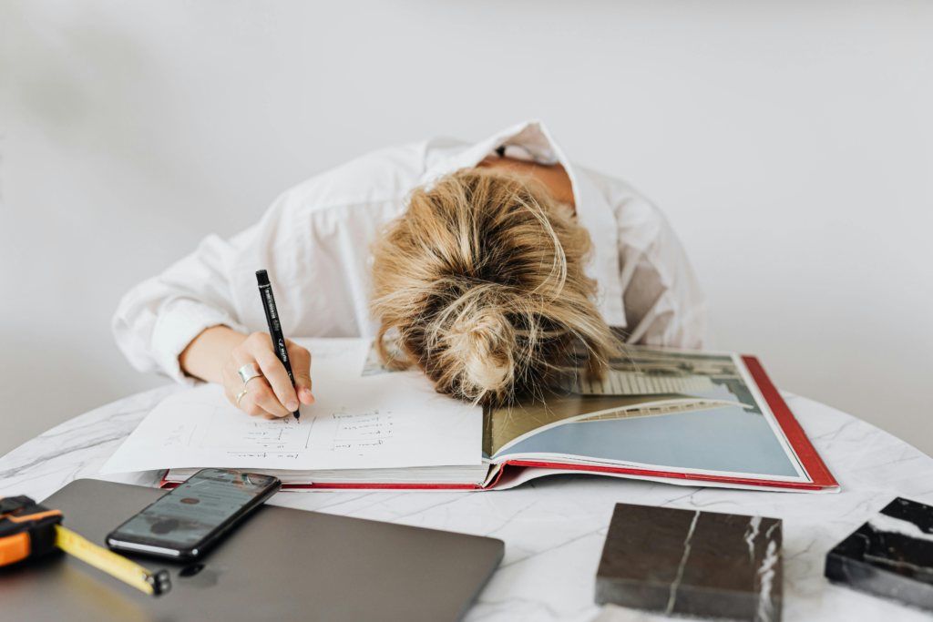 A tired teacher rests her head on an open book at her desk, indicating stress or fatigue.