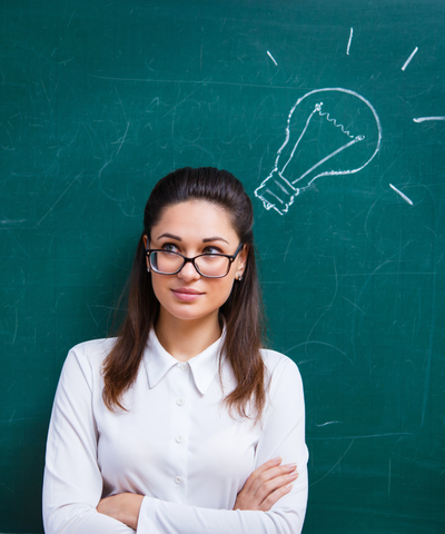 A teacher stands in front of a chalkboard with a chalk drawing of a lightbulb by her head signifying all the new ideas she received from classroom management coaching. Her arms are crossed and she is smiling confidently, secure in the knowledge that she can easily control her class.