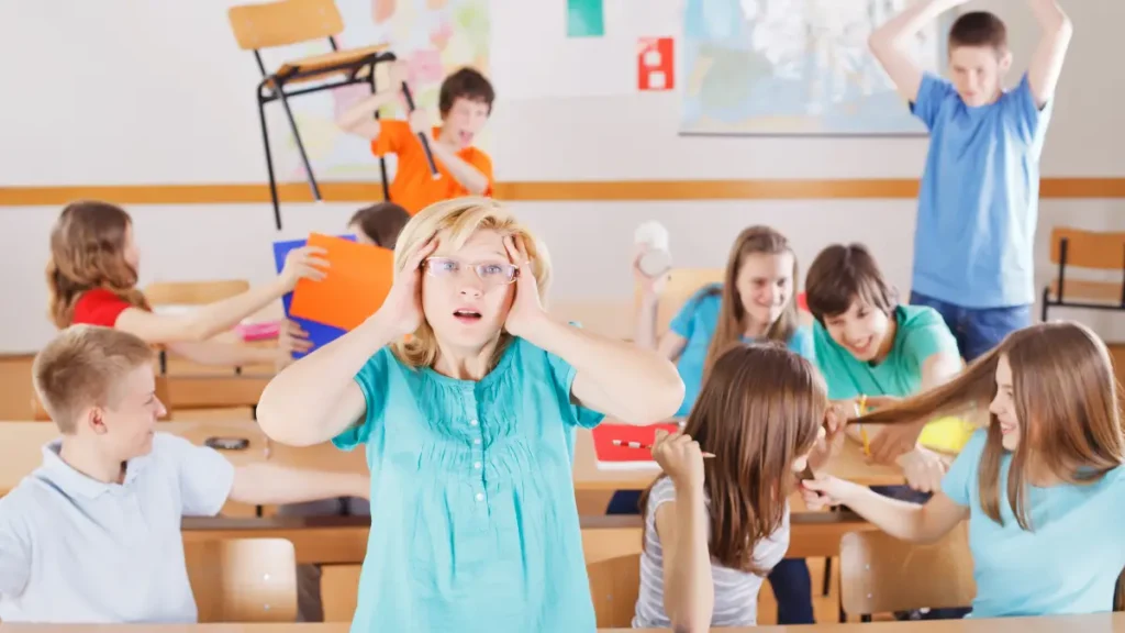 A blonde teacher wearing a teal shirt puts her hands to the sides of her heads and widens her eyes in dismay at the chaos behind her. Students are raising chairs above their heads, throwing paper, and pulling each other's hair, showing a classroom transition strategy that is not working.