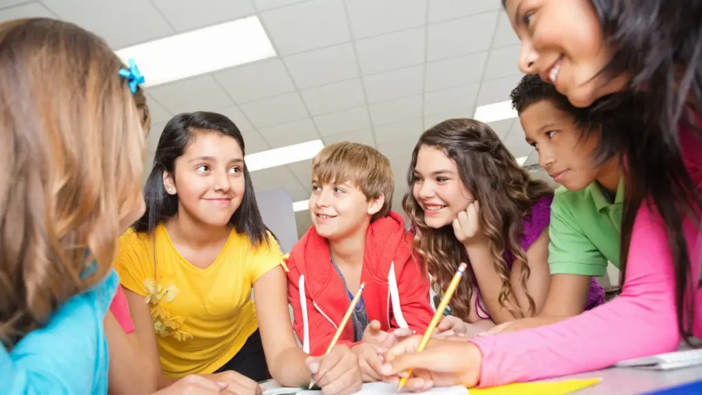 A group of older elementary students or middle schoolers lean over to work on their independent work while looking at a peer student in a yellow shirt who smiles confidently.
