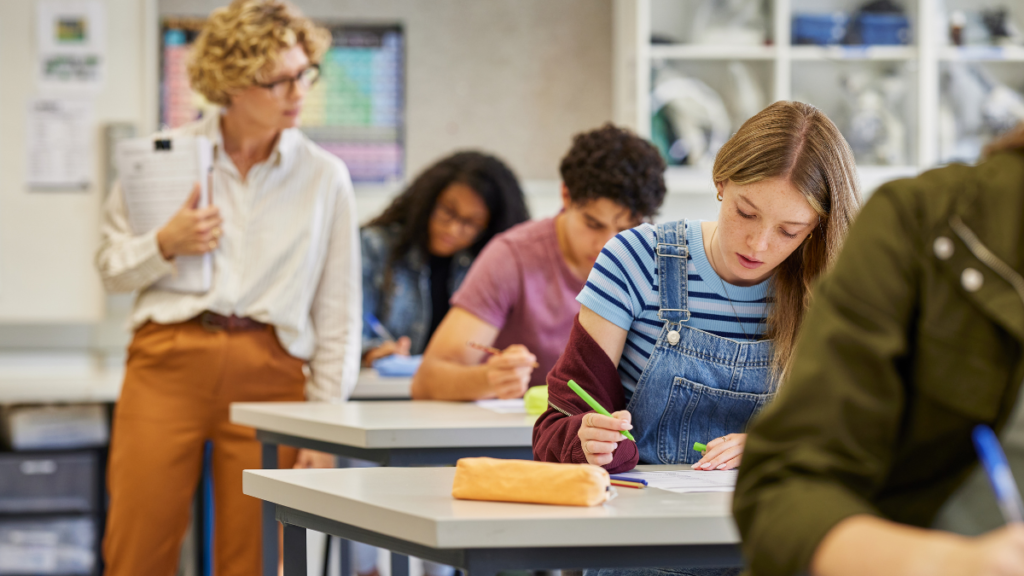 A teacher walks around and checks on a class of older students. There are four students visible, while there is only one teacher, showing that the teacher cannot help every student at once.