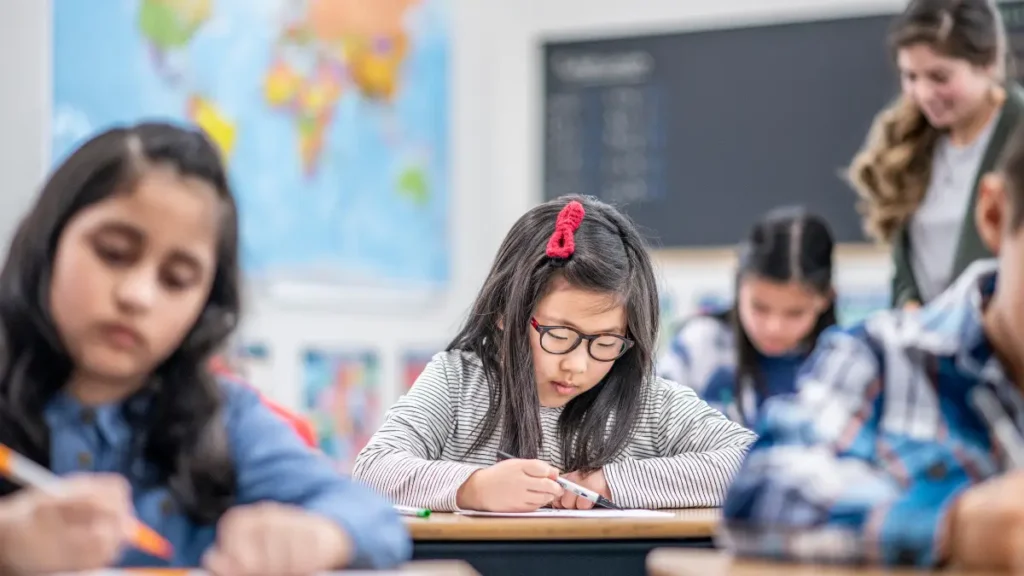 A fourth grade student works at her desk while the teacher hovers in the background, standing by a student who won't do their work.