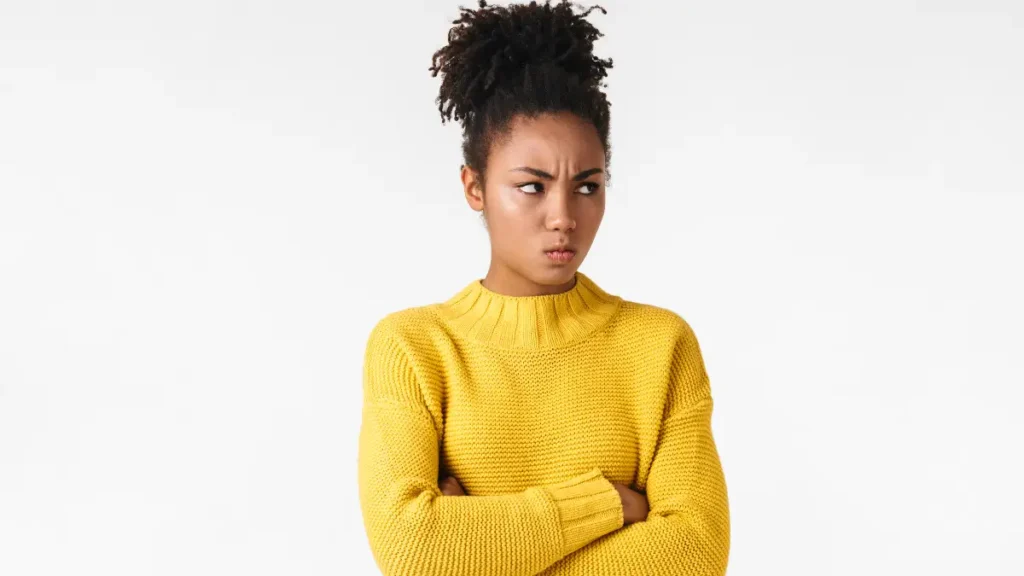 A young teen wearing a yellow sweater furrows her brows and scowls. Her arms are crossed and she looks to the side, showing that she feels defiant or wants to avoid doing work.