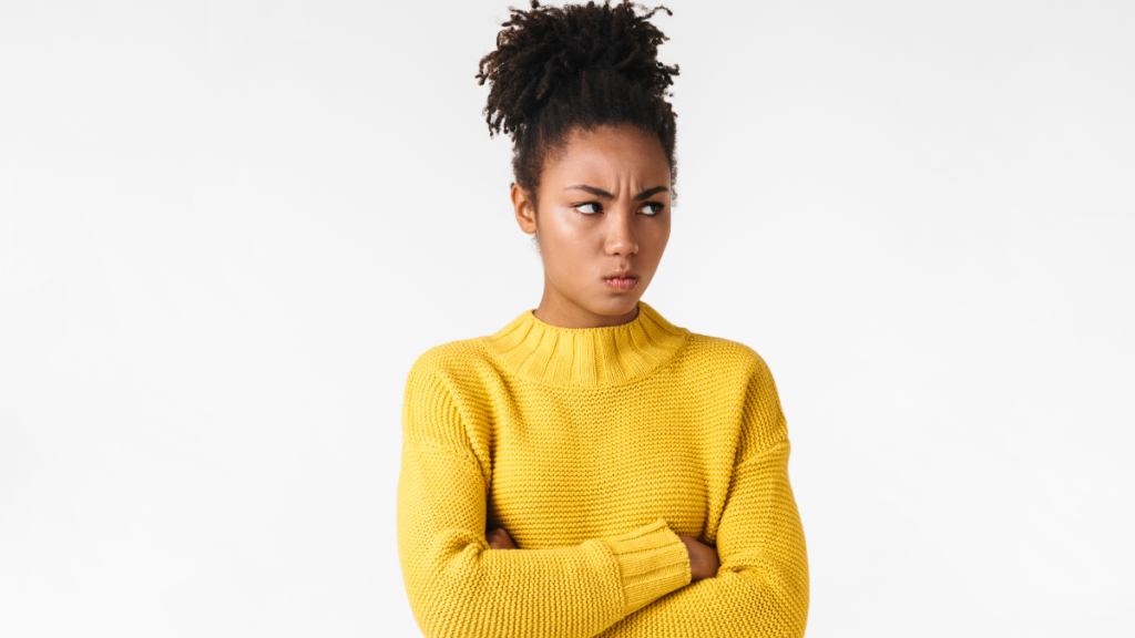 A young teen wearing a yellow sweater furrows her brows and scowls. Her arms are crossed and she looks to the side, showing that she feels defiant or wants to avoid doing work.