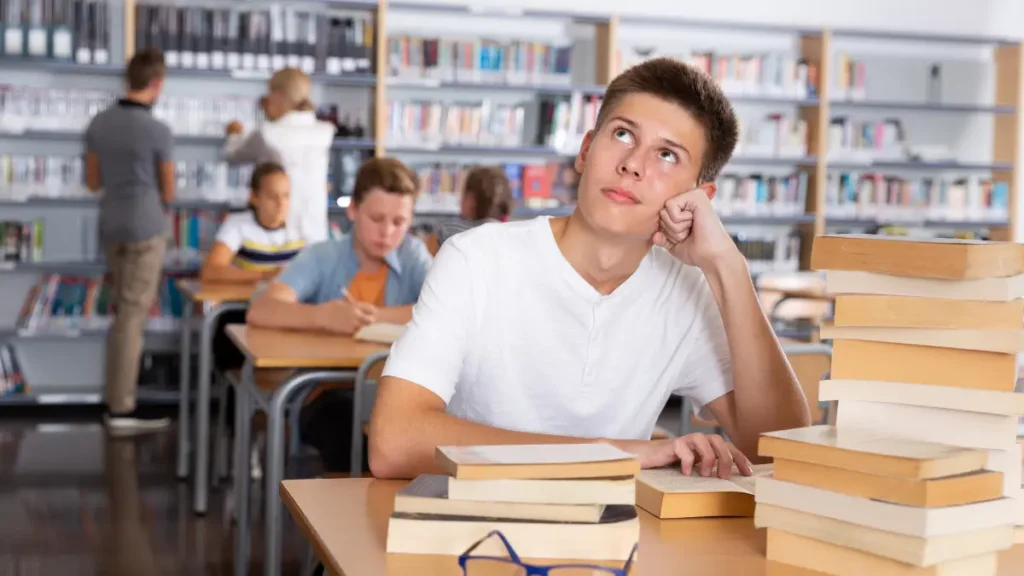 A male student in a white t-shirt sits at a desk behind stacks of books. His eyes gaze up and left, indicating that he is distracted by his environment or his own thoughts, which prevents him from doing his work.