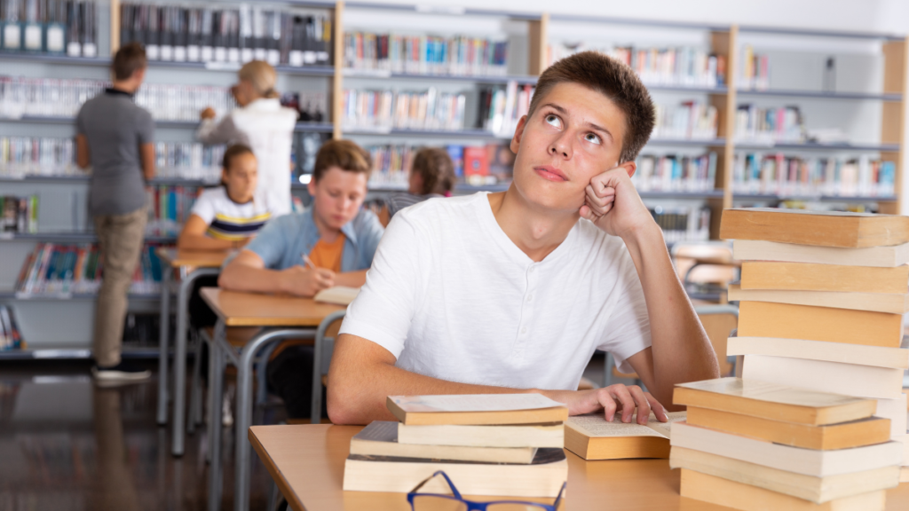 A male student in a white t-shirt sits at a desk behind stacks of books. His eyes gaze up and left, indicating that he is distracted by his environment or his own thoughts, which prevents him from doing his work.