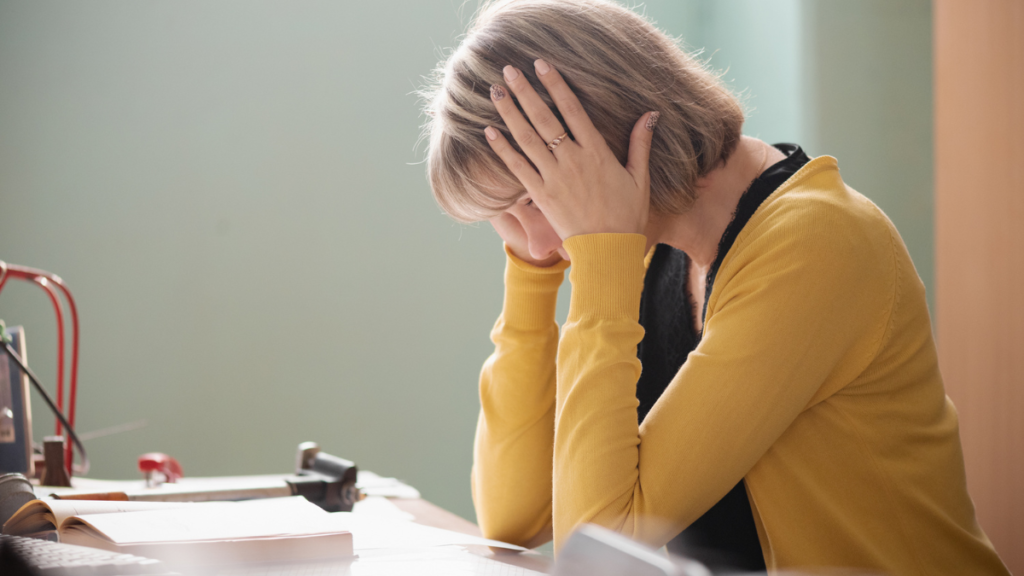 A teacher with short blonde hair and a yellow sweater sits at her desk in profile and holds her head in her hands, indicating burnout and exhaustion from chasing students who won't do their work.