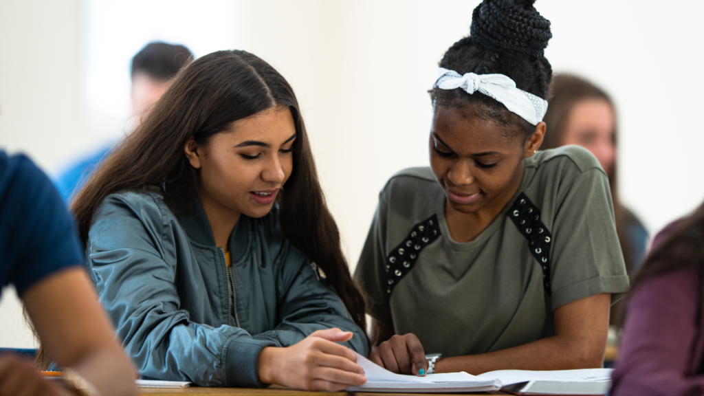 Two students sit side by side looking at the same paper. One student points her finger at the paper, indicating something as she explains a concept to her classmate.