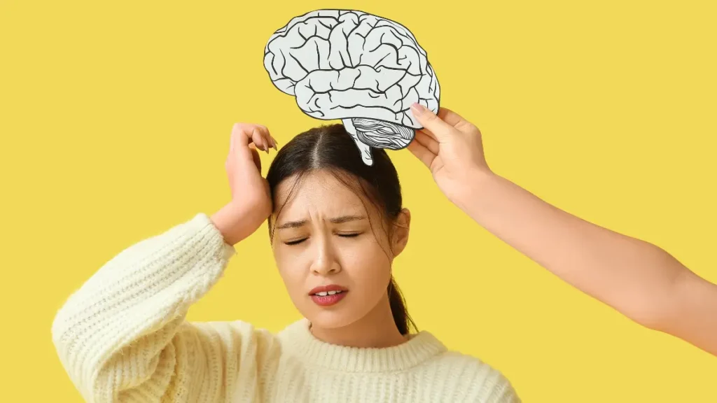 An older student grimaces and holds her hand to her forehead while a hand off-camera holds a paper brain above her head, indicating that her brain needs a break.