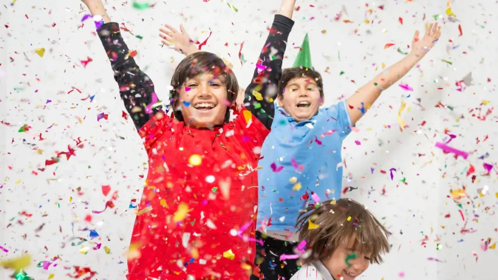 Three older elementary students throw their hands up and grin as they are showered by confetti, showing enthusiasm and excitement for a short physical activity break in their classroom.