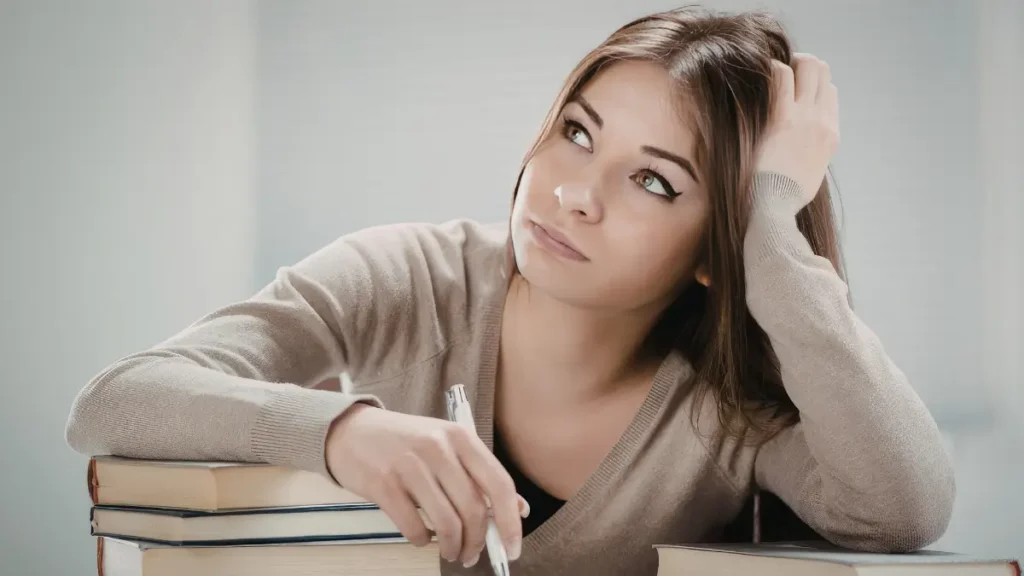 A middle school student with her arm laid on a stack of books rests her head in her hand as her eyes stare off the edge of the photo. She is clearly distracted and disengaged from the task.