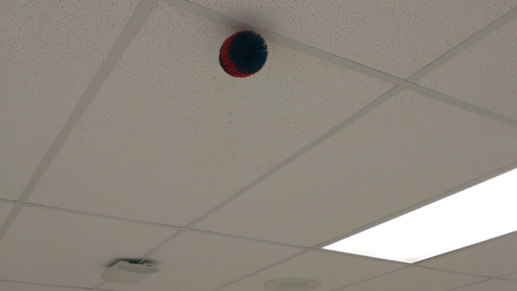 A stringy (Koosh) ball hangs unsupported from a drop ceiling, where the stringy tendrils wedged into the ceiling support pieces after getting tossed there on accident during a class discussion.