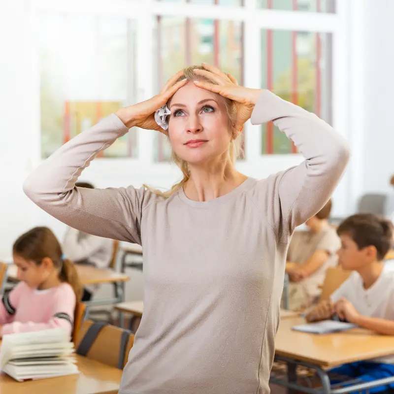 A blonde teacher holds her hands to her head and gazes into the distance while a class of students works behind her. The teacher's expression is one of annoyance and frustration, showing her difficulty with classroom management and a desire for a better classroom system.