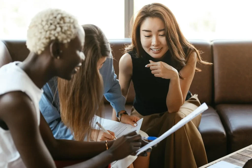 Three women sit on cushions looking over paperwork such as a student's report card or sample work during a parent teacher conference