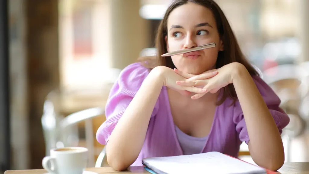 An older student balances a pen between her mouth and nose while staring out a window with a blank notebook in front of her, showing that it might be time to use a brain break