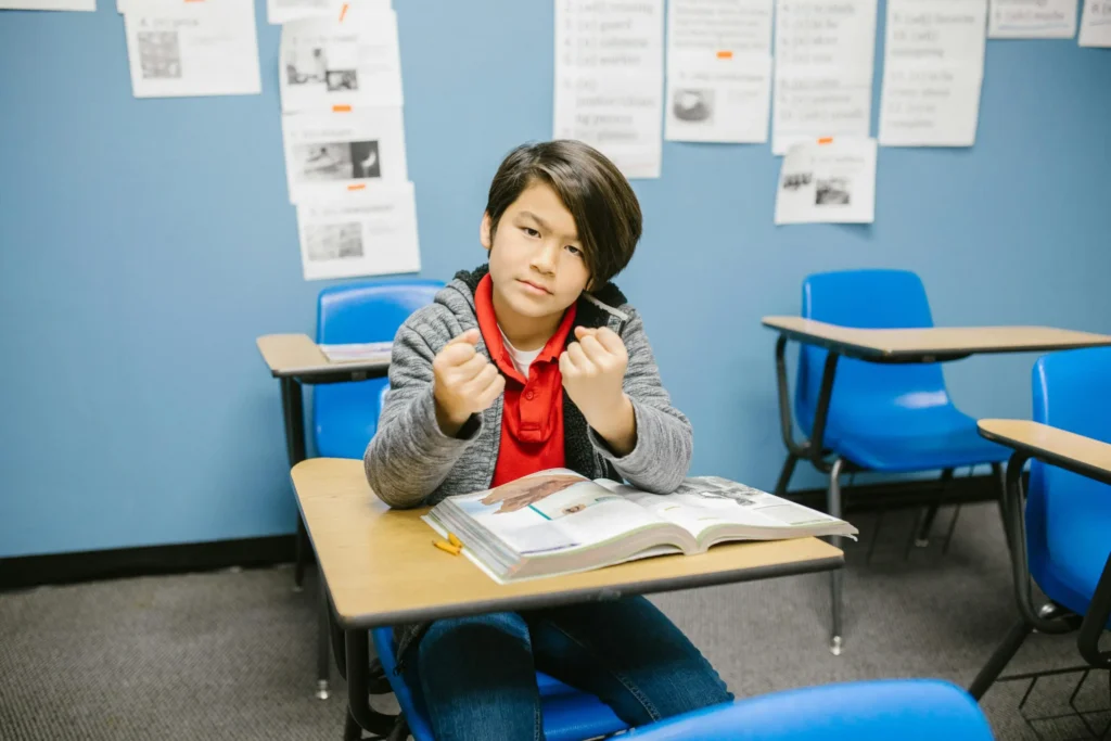 An upper elementary boy sits at a desk with a book open in front of him. His fists are clenched and his eyes are narrowed.