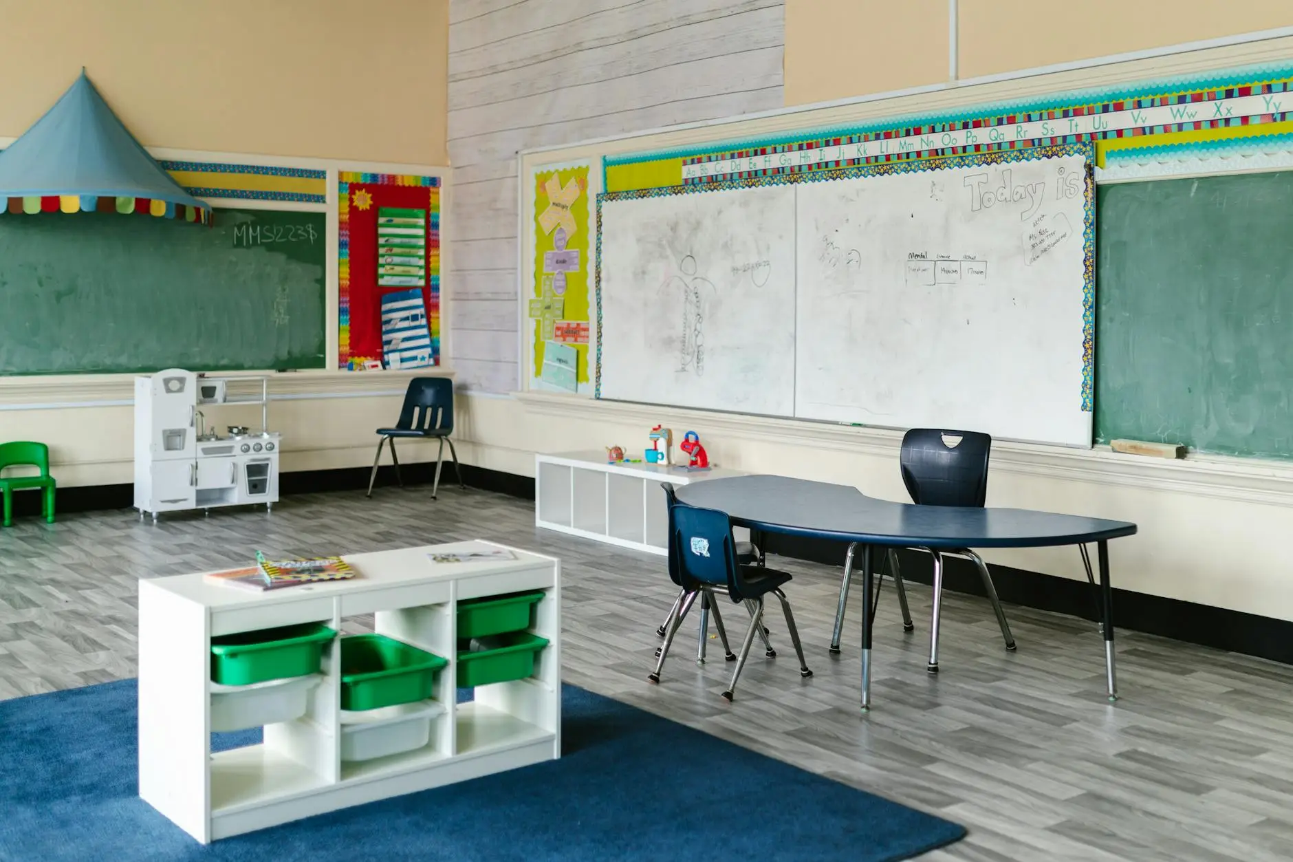 A clean classroom with a whiteboard, blue table, chairs, and bins. Everything is neat and organized, ready to go before the first day of school.