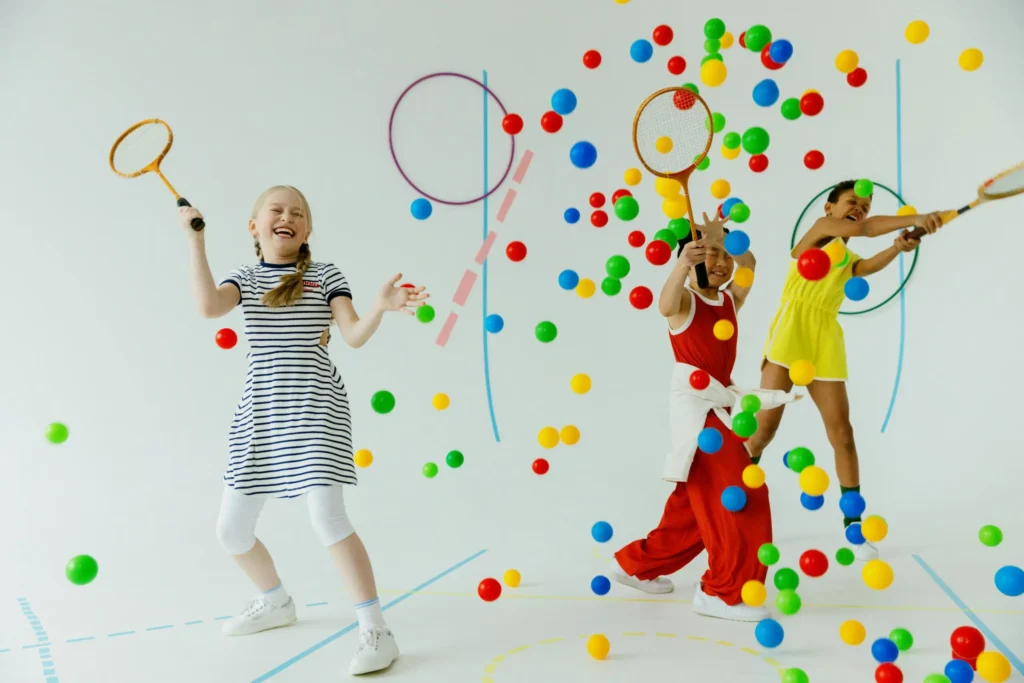 Children playing indoors with rackets and colorful balls, showcasing joy and diversity.