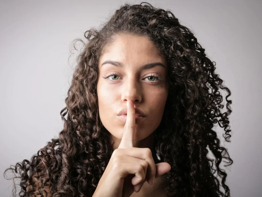 A woman with curly brown hair and green eyes holds her finger to her lips in a "shh" gesture