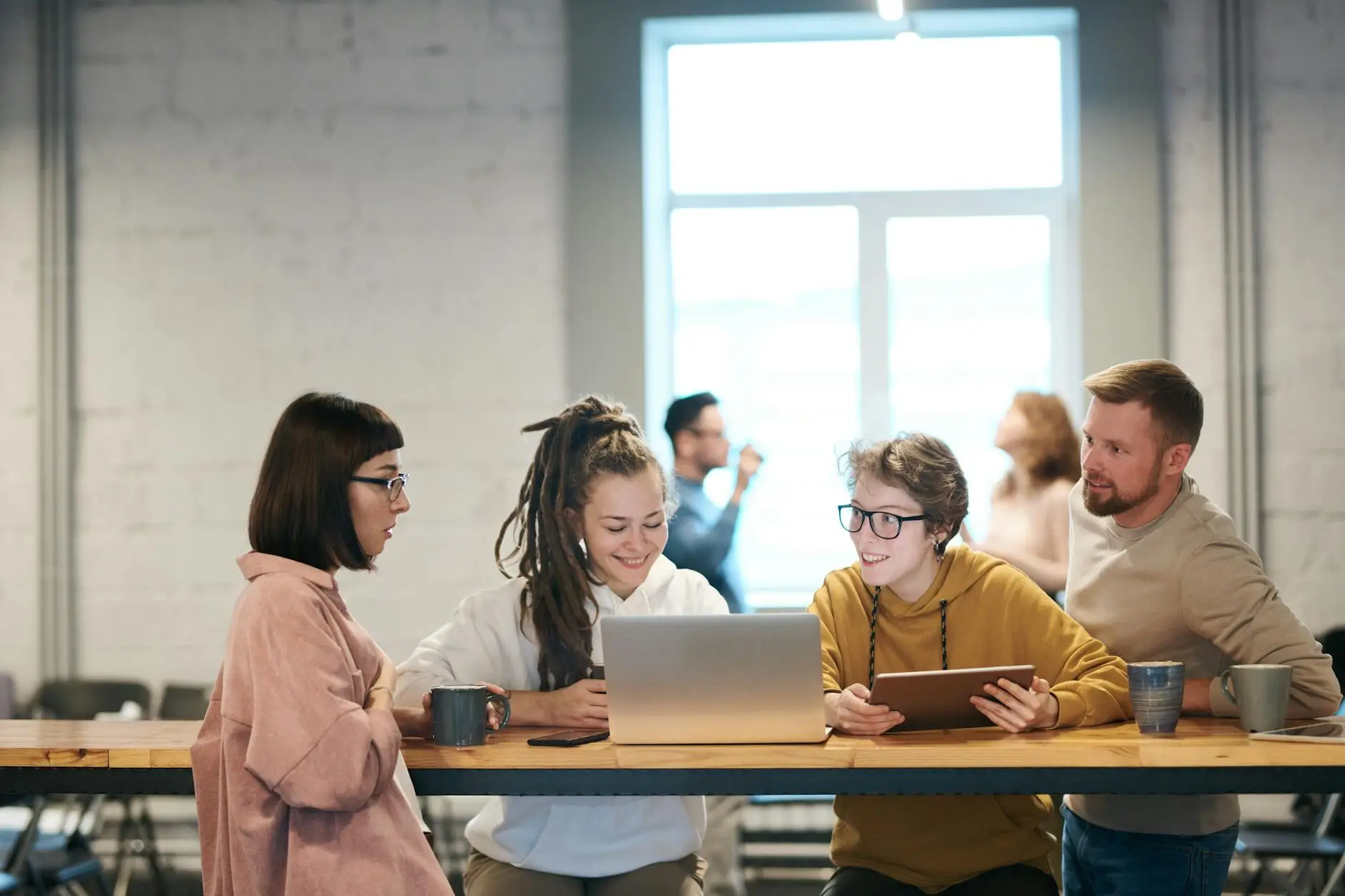 Four people smiling and gathered around a computer, sharing ideas over coffee at a wooden table.