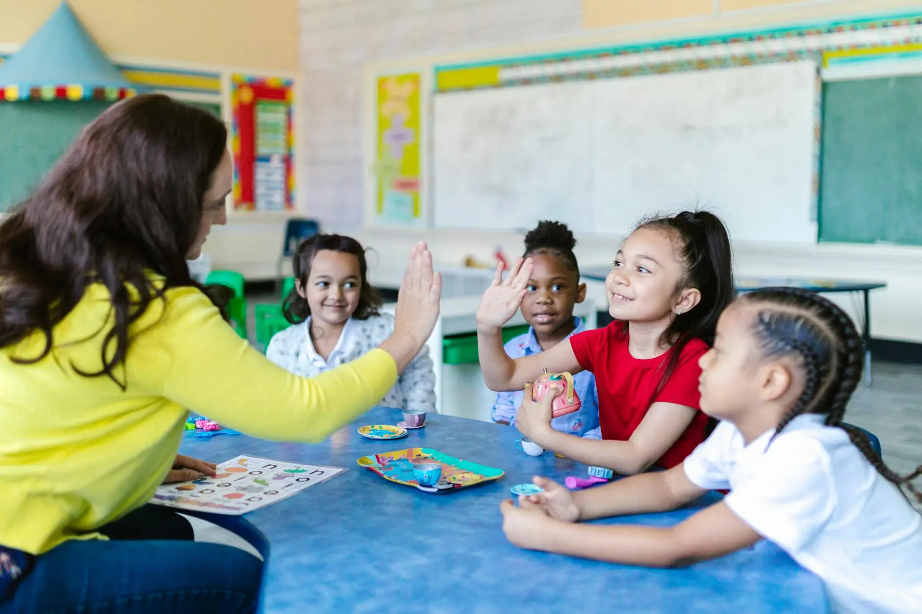 A teacher sits behind a blue table while four young students look on. The teacher gives one student a high-five.