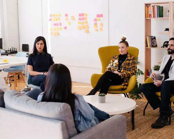Several teachers sit in a circle in a teacher's lounge or office. They are collaborating and talking with each other in this supportive environment.