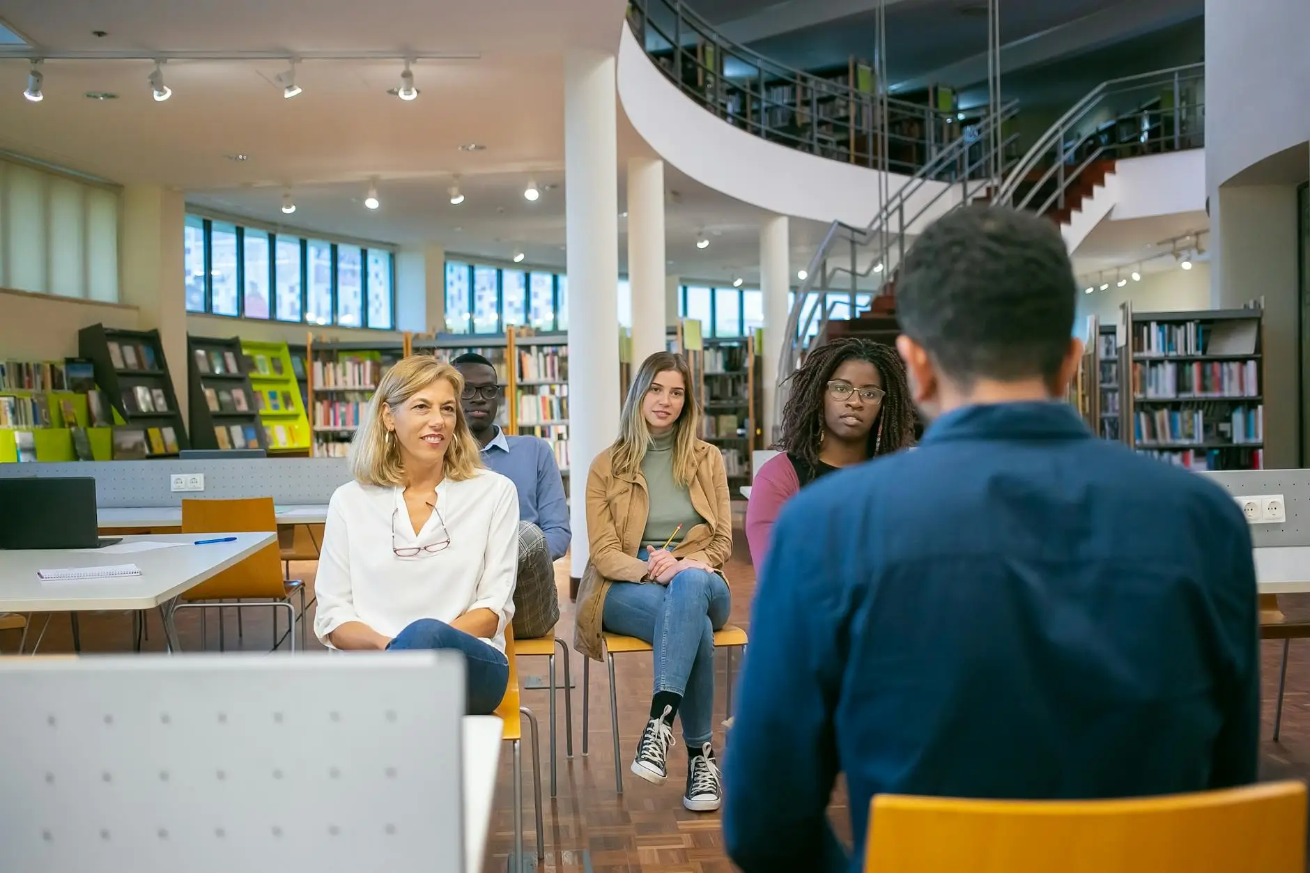 Several parents sit in a library, listening to a teacher introduce parent-teacher conferences. The parents are smiling, showing confidence in the teacher's methods and preparation.
