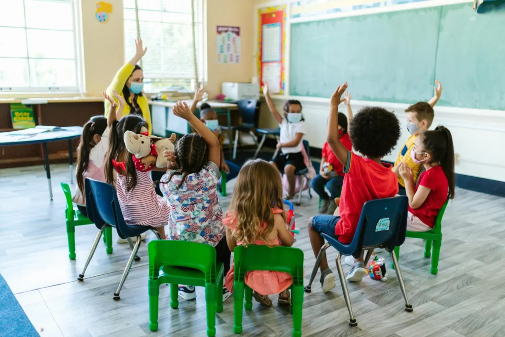 Diverse group of young children in a classroom, raising hands, participating in a class meeting led by their teacher. Bright and engaging learning environment.