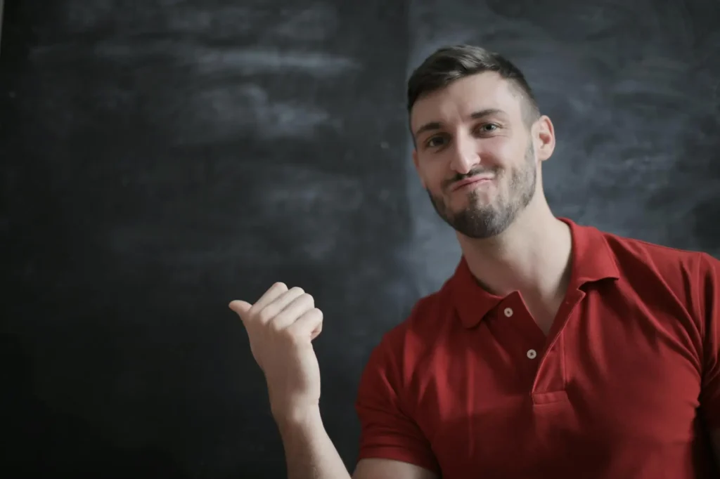 A male teacher sits in front of a blackboard and motions with his thumb to the side, his facial expression and gesture indicating permission to use the bathroom without saying a word.