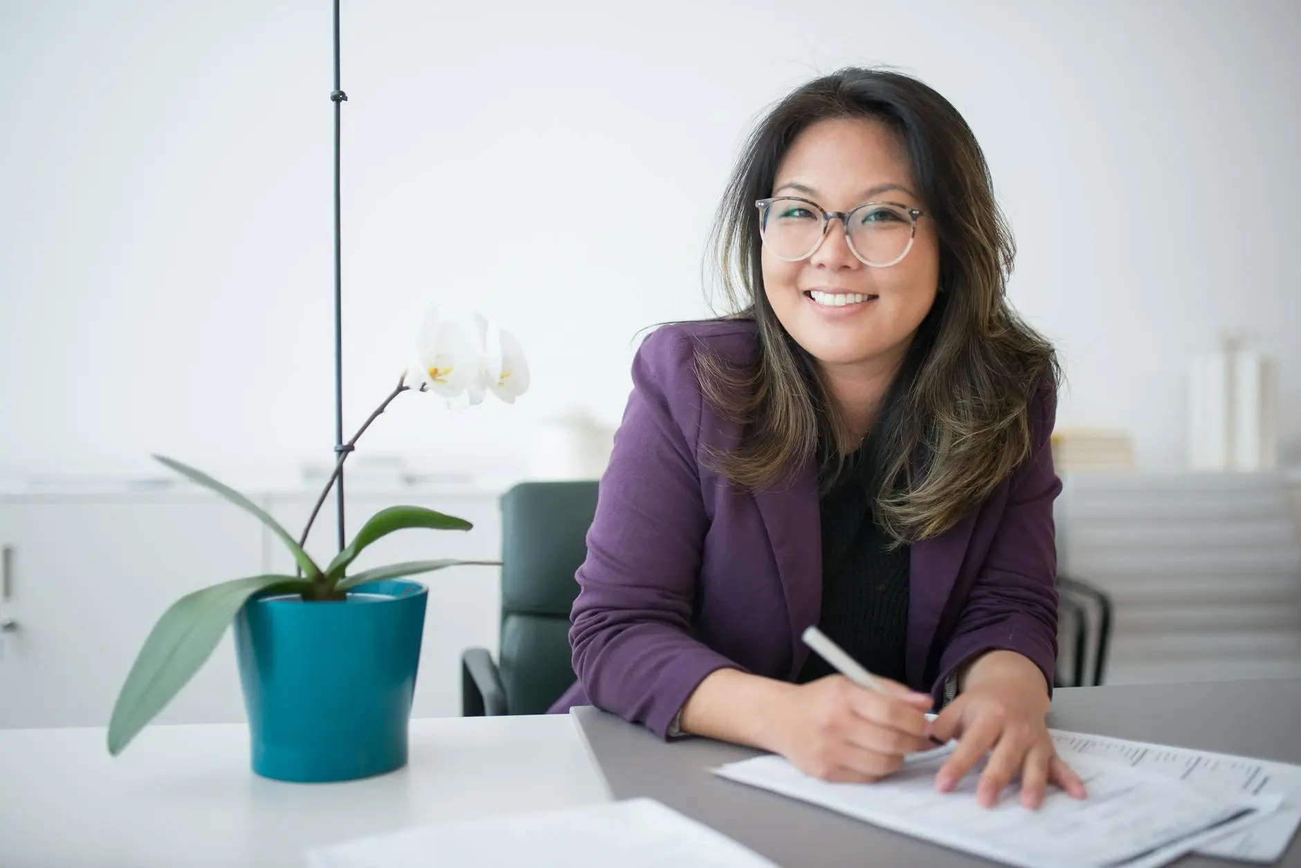 A woman in a purple blazer jacket sits at a desk with a pen ready. She is smiling, indicating confidence and eagerness to write her classroom management plan.