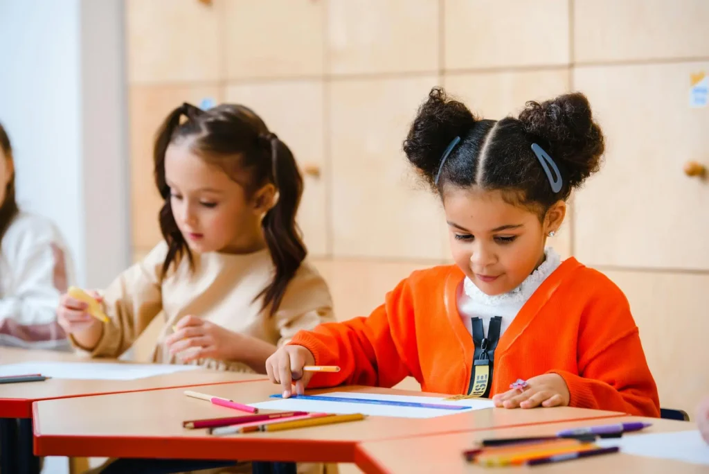 A young girl in a red sweater sits at her desk and draws beside her friend, showing an easy time-filler activity for young students