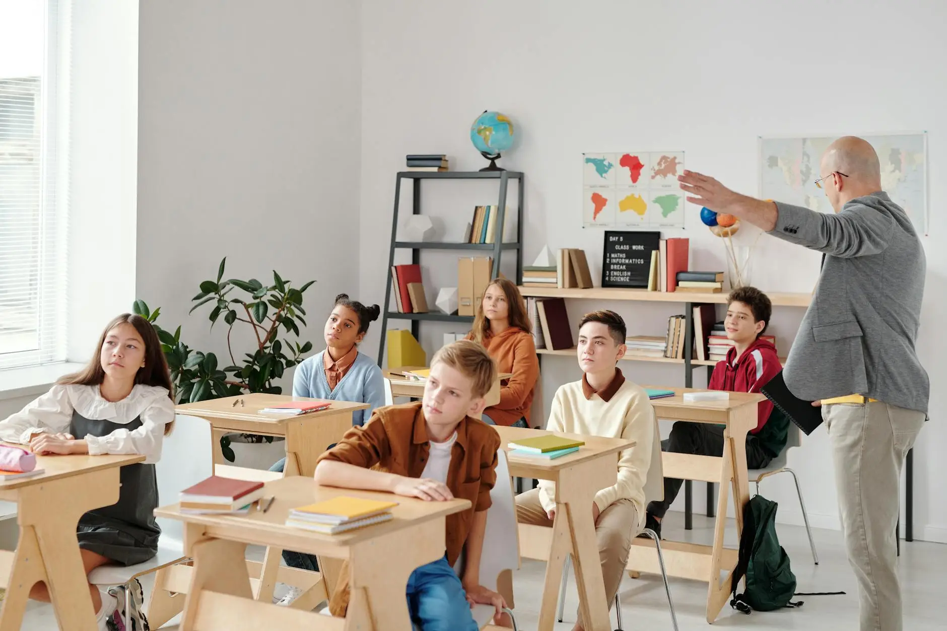A teacher gestures to the front of the room while students look attentively where he is indicating. The students are sitting obediently in orderly rows, showing that the teacher has a strong grasp of classroom management.