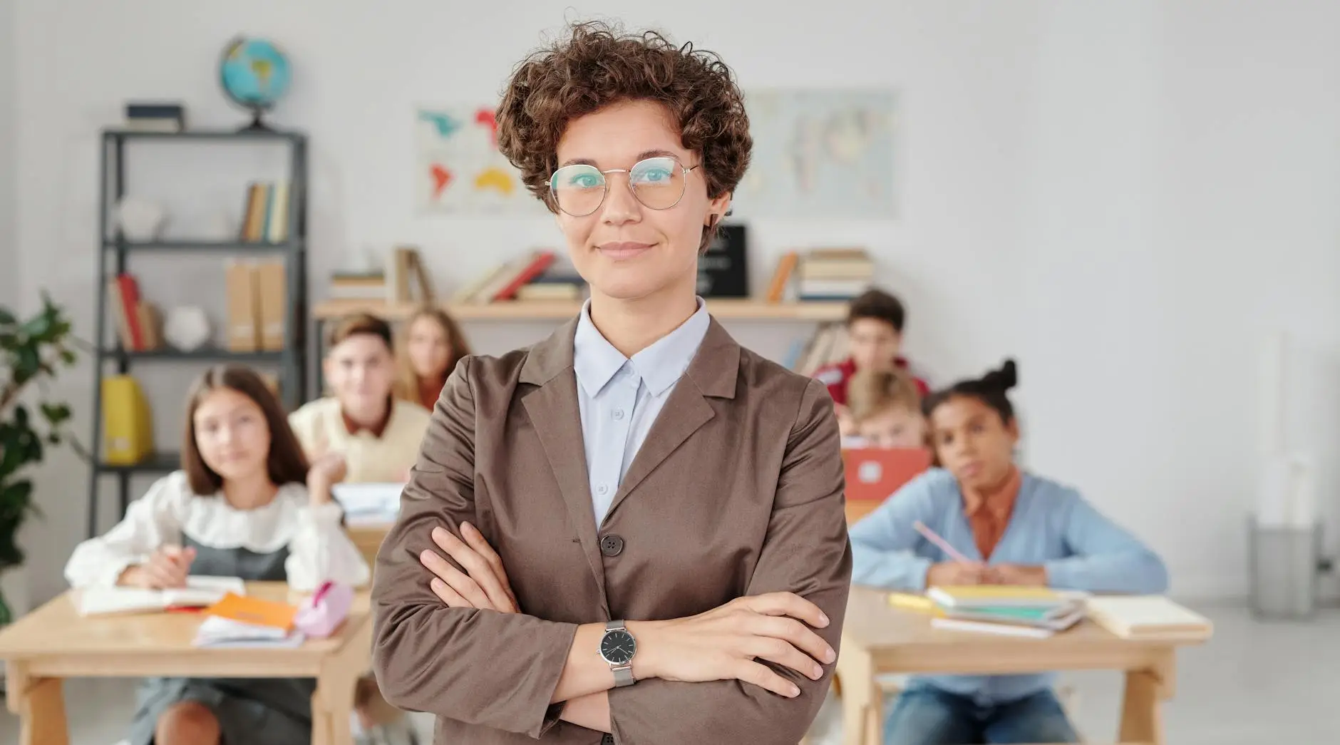A teacher in a brown blazer crosses her arms and smiles confidently as her students wait patiently behind her