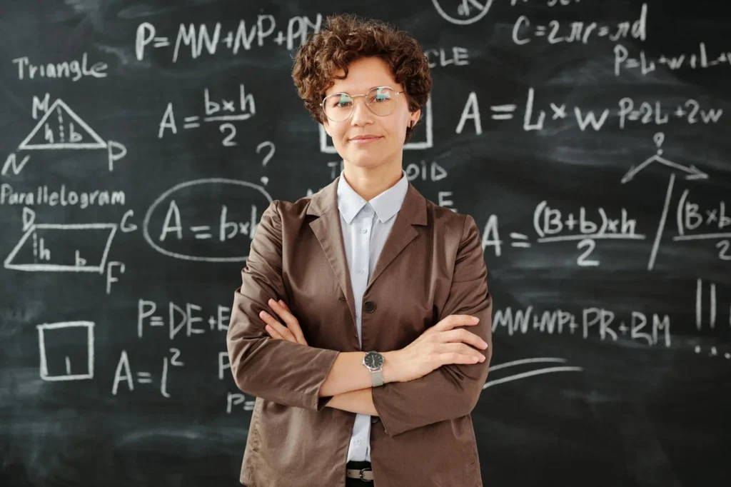 A teacher in a brown jacket stands in front of a blackboard covered in math equations. Her arms are folded and her expression is patient, showing a willingness to wait for student misbehavior to end before she smiles and continues the lesson.