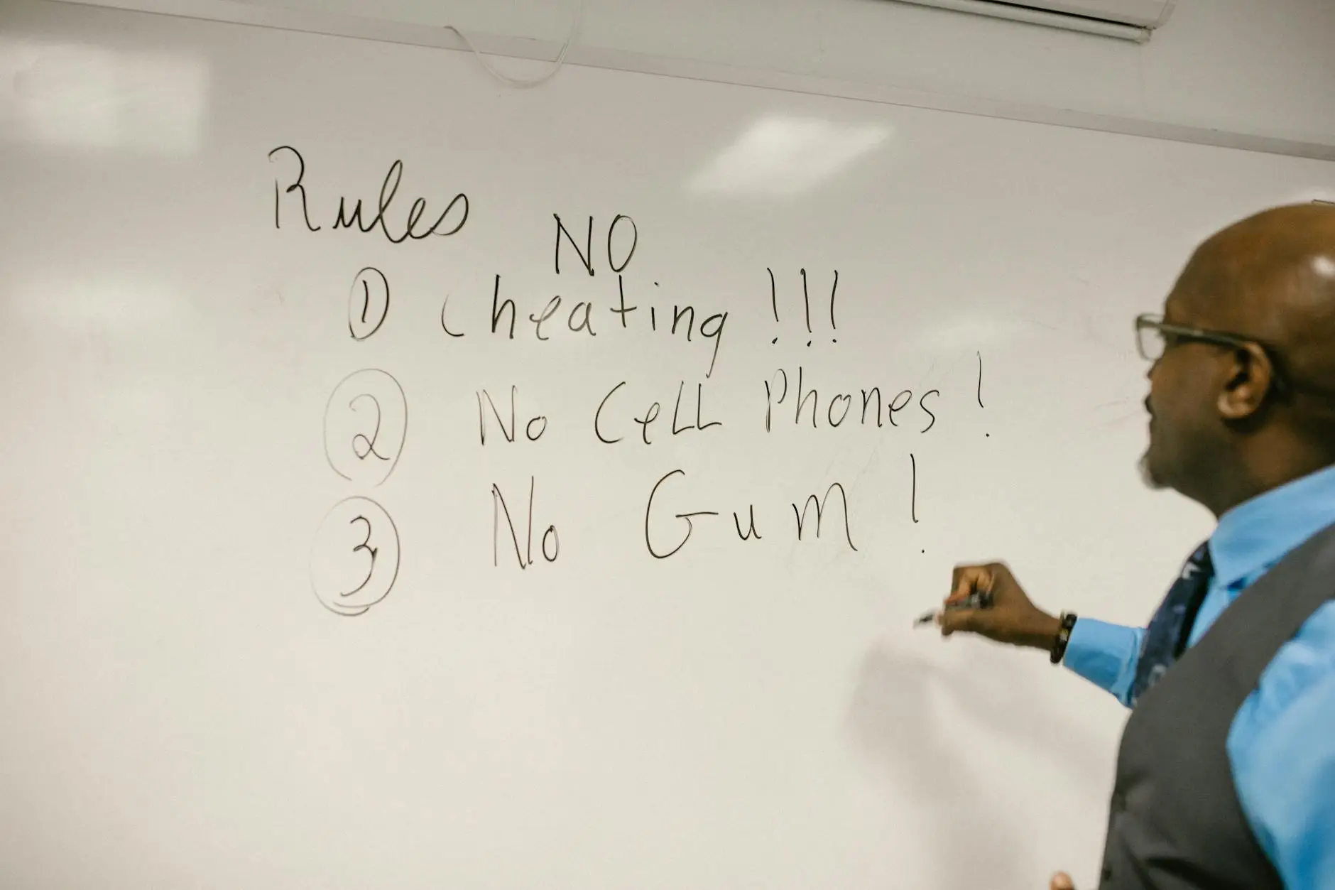 A teacher writes classroom rules on the board, including no cheating, no cell phones, and no gum