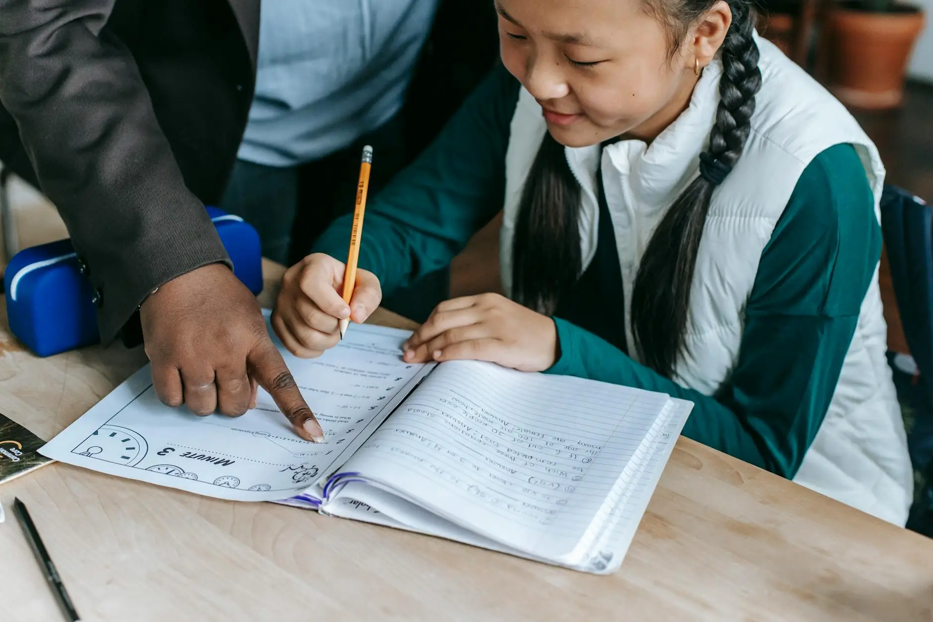 A teacher leans over a student's desk and points to a part on the student's worksheet. The student holds a pencil and smiles, ready to continue working.