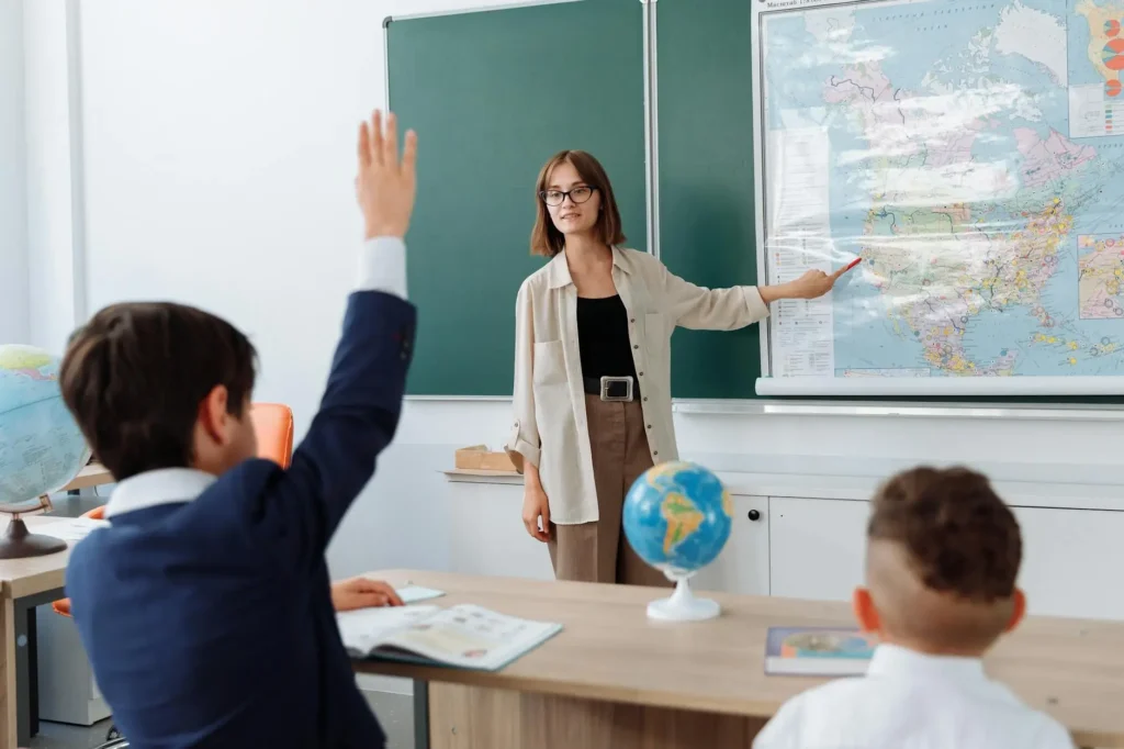 A teacher points to a map on the wall while a student raises his hand. The teacher looks hesitant, unsure if the student is raising his hand to answer a question or to ask to use the bathroom.