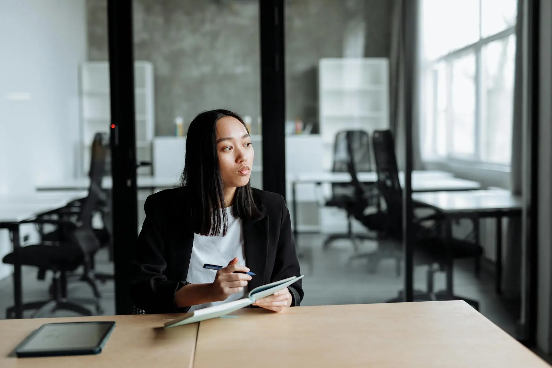 A teacher with long black hair sits at a clean desk in a conference room staring into the distance. She is holding a notebook and pen and is taking notes in preparation for parent-teacher conferences
