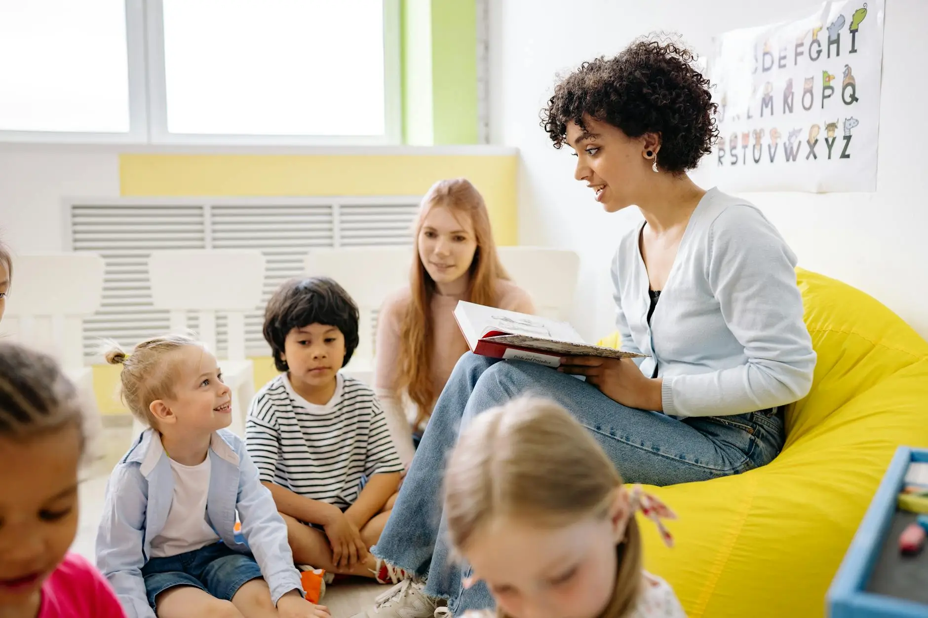 A young teacher sits in a yellow beanbag chair while students sit on the floor and listen attentively