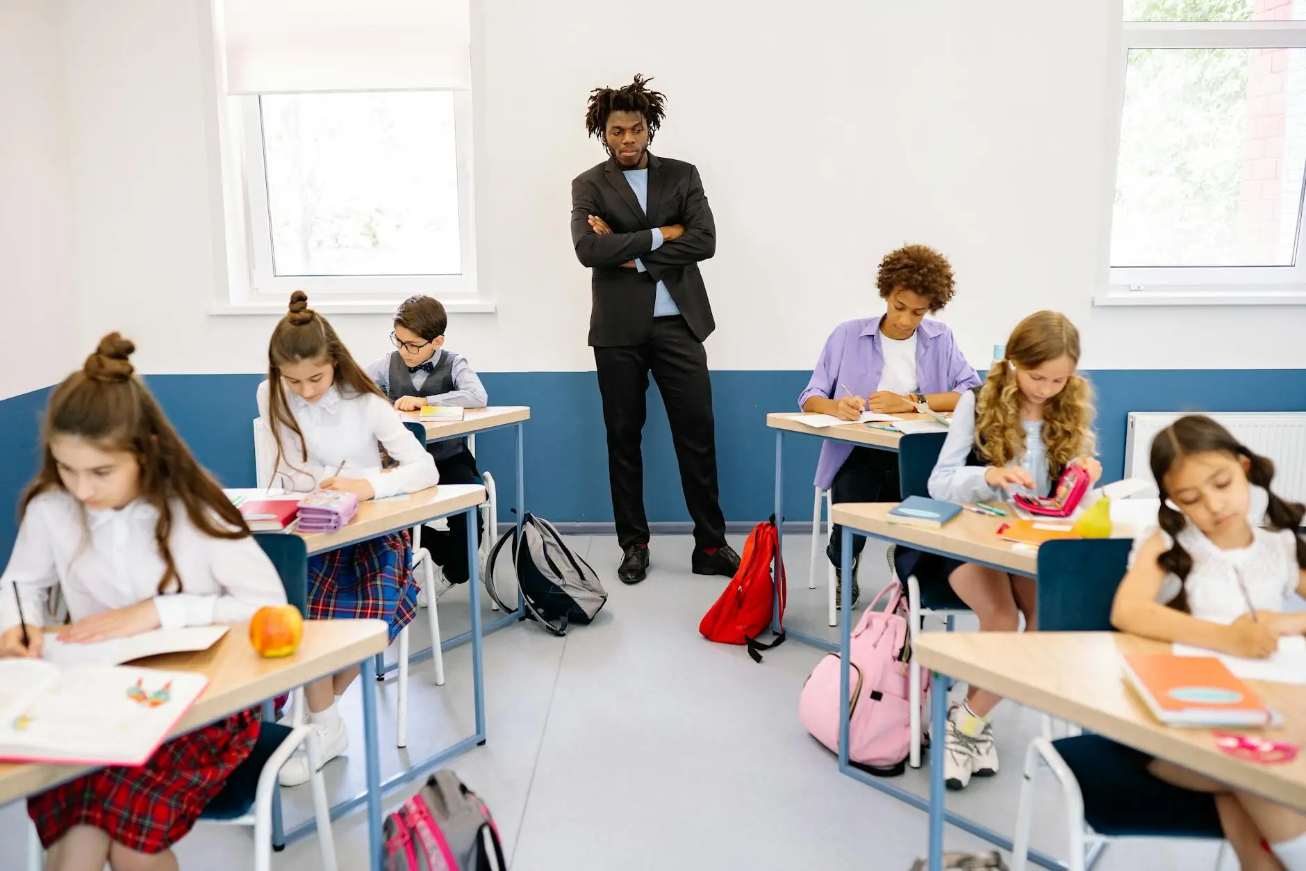 Students sit calmly and complete their work at their desks while their teacher looks on.
