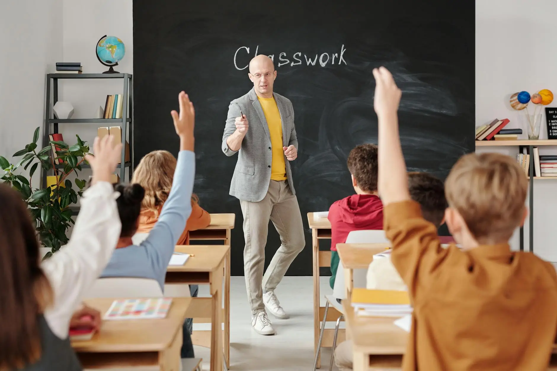 A teacher in a gray jacket and yellow shirt stands at the front of a room in the process of calling on a student. Several students have their hands raised, but every student is seated and paying attention to the lesson. They are not disruptive or wild, showing that the teacher has effective classroom management strategies in place.