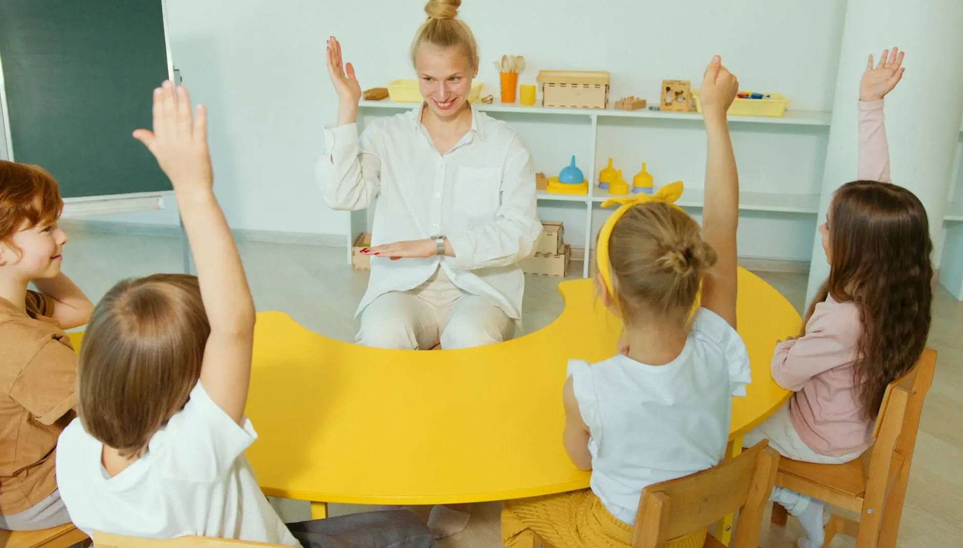 A blonde teacher sits at a yellow table with young students. The teacher is modeling raising her hand, and the students are following her example.