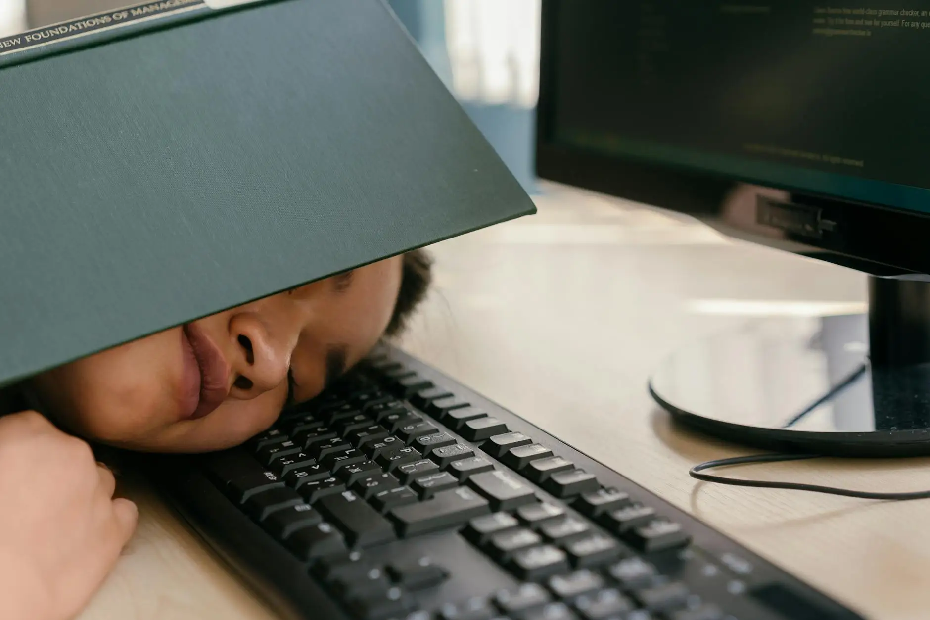 A student lays their head on a computer keyboard with an open book over their head, taking a nap due to boredom.