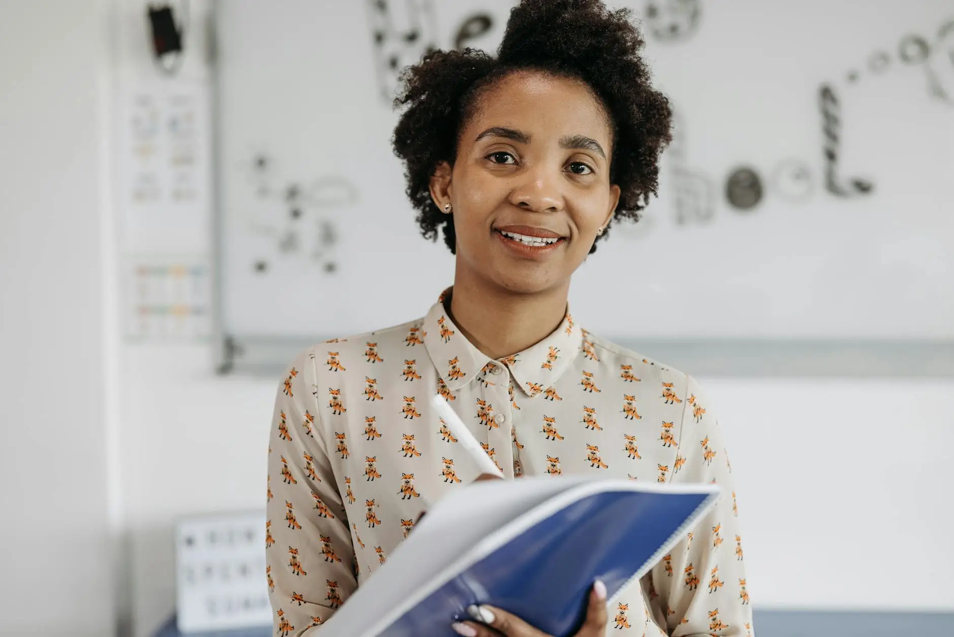 A teacher with short curly hair wearing a fox-patterned blouse smiles as she prepares to write in a blue notebook