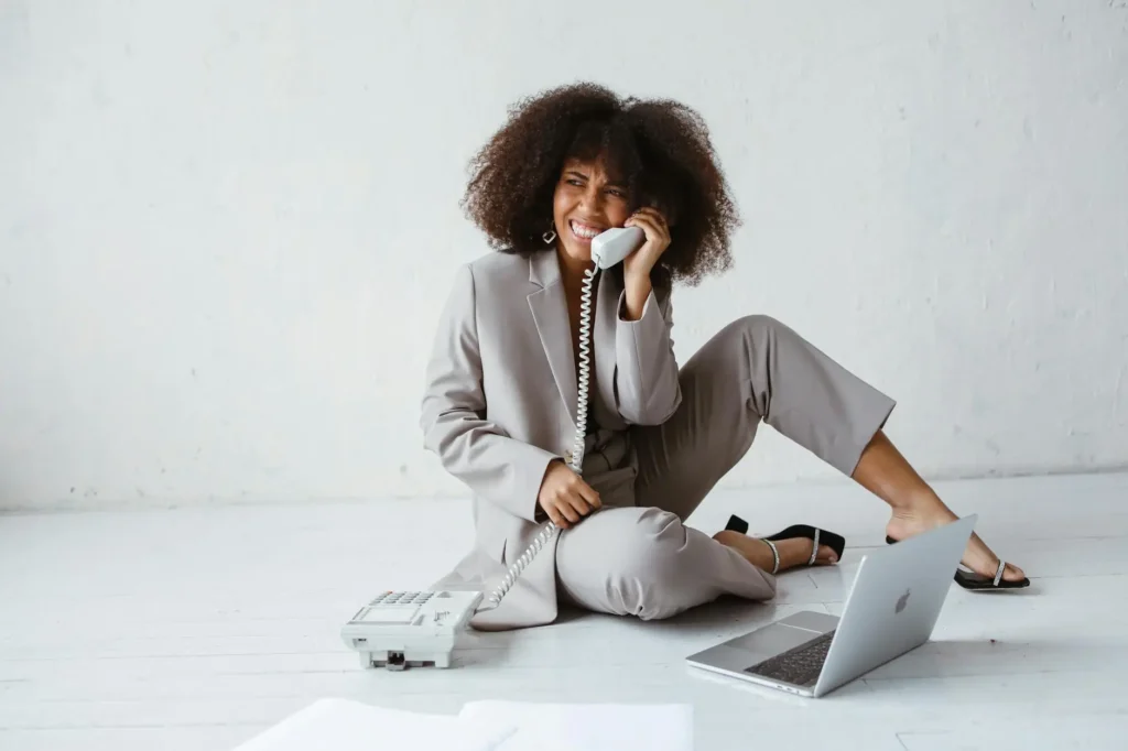 A teacher in a gray suit sits on the floor beside a landline telephone. Beside her is a notebook and an open laptop, showing that she is well-prepared as she contacts the parents of one of her students