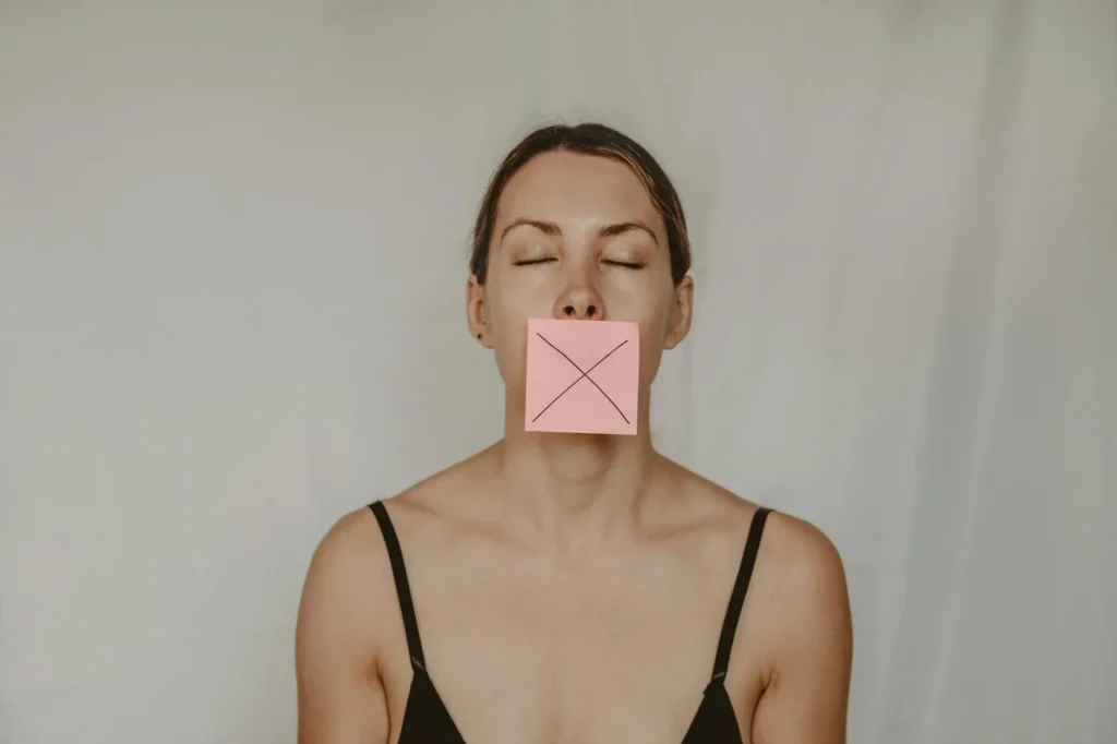 A young woman in a black tank top has a pink sticky note with a black X across her mouth, showing that she is not talking