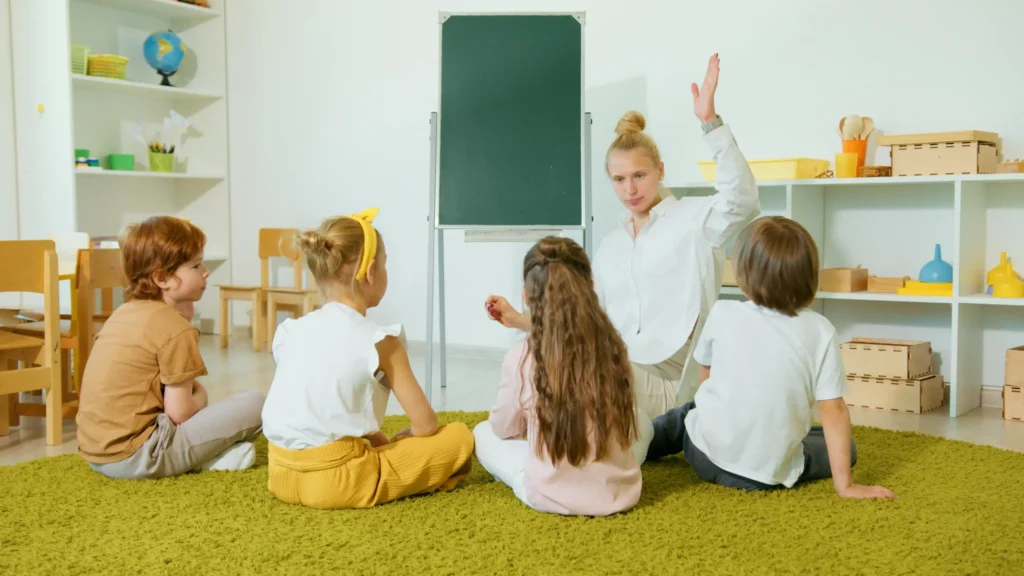 Students sit on the floor in front of a teacher, who models raising her hand to ask a question