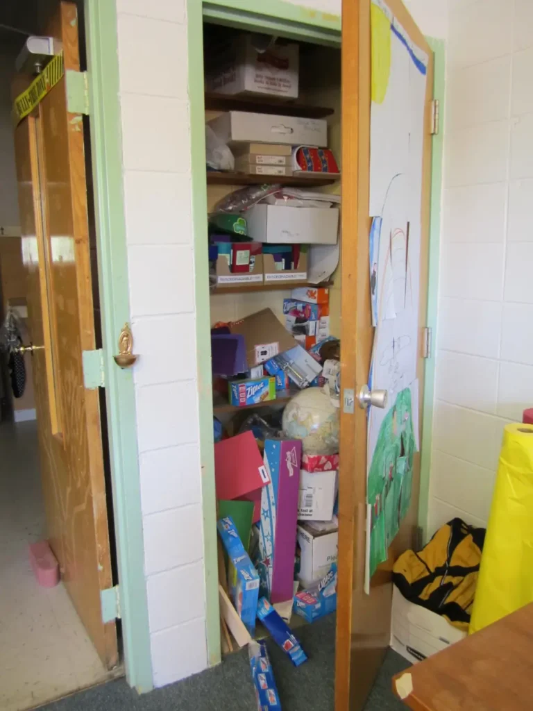 A messy closet during classroom setup with boxes, bags, and school supplies spilling out the half-open door