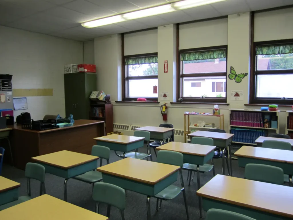 A classroom with desks in orderly rows in front of a bookshelf full of textbooks and next to a teacher's desk